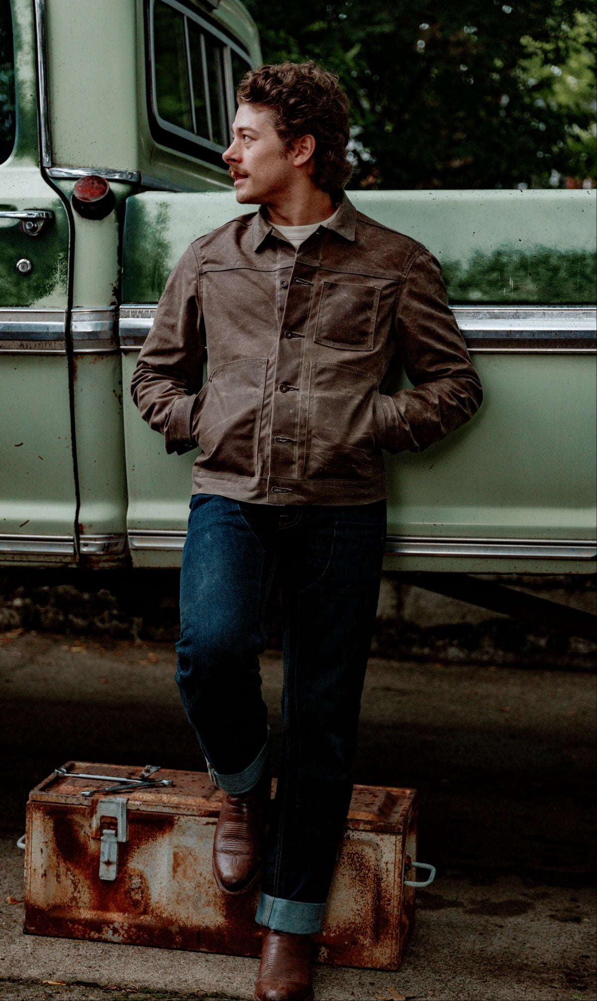 A man wearing a brown corduroy utility jacket, blue jeans, and brown boots stands in front of a vintage light green truck, leaning against it and resting one foot on a rusted metal trunk.